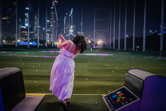 Woman in white dress swings golf club at Topgolf Dubai with illuminated city skyline and vibrant lights in the background