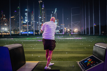 Golfer in white shirt and shorts swings club at Topgolf Dubai with illuminated city skyline in the background at night