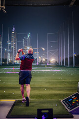 Obraz premium Man in colorful attire swings golf club at Topgolf Dubai, with city skyline illuminated at night in the background