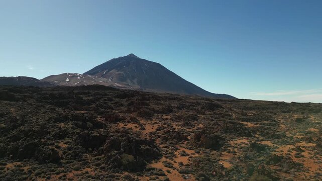 A stark volcanic landscape unfolds under a clear blue sky, leading to a majestic, snow-dusted mountain peak in the distance.