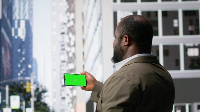 Horizontal chroma key screen held by black man near pedestrians walking with steady motion through the cityscape. Urban crowd diversity, routine commute and action of modern city life. Camera B.