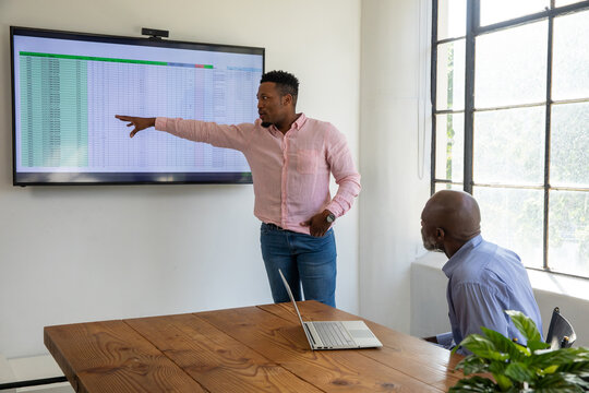 African American men presenting and pointing at spreadsheet on monitor in meeting room with laptop