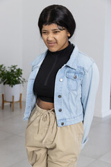African American woman standing squinting hands behind back in studio with plant denim jacket pants