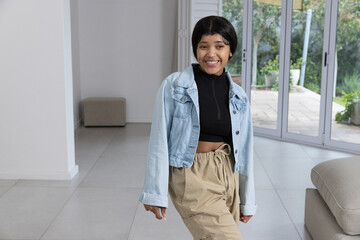 Woman smiling and wearing denim jacket and drawstring pants in living room near sliding glass doors