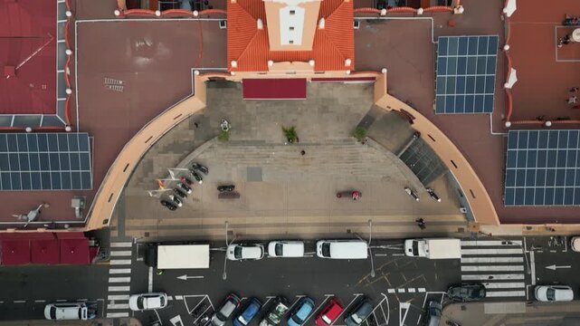 Aerial view shows a building with solar panels, a plaza with a few people, and a street with parked cars and a crosswalk.