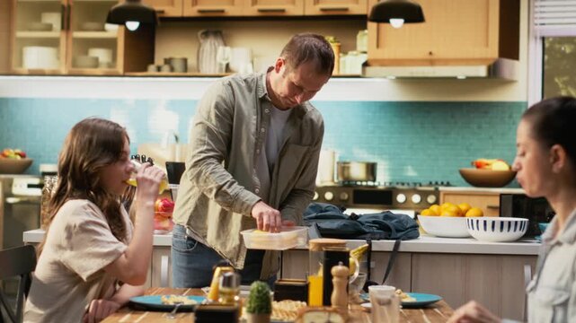 Father packing school lunch for his pre-teen daughter and preparing backpack, family members enjoying breakfast together during cozy morning in the kitchen. Meal prep school bag. Camera B.