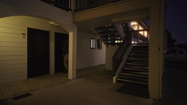 Adult man walking alone through outdoor motel corridor at night, carrying bag and returning to room. Travel lifestyle scene, road trip stop, temporary accommodation, solitude, evening routine.