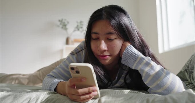 Young woman on bed using smartphone in soft morning window light ,Asian teenage lies on a neatly made bed, focused on a smartphone. Soft morning light from a window illuminates her striped sweater