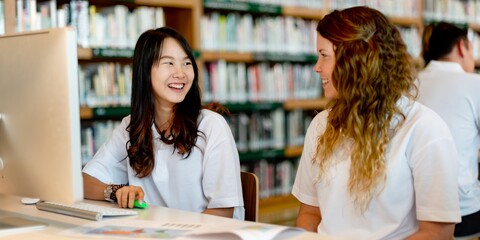 Diverse students of Asian and Caucasian women studying together, using computer in a library. Students collaborating using technology. Diverse students in library setting. High school education.