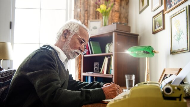 Elderly man writing at a desk with a typewriter. Focused elderly man, glasses, white hair. Cozy room, typewriter, writing. Elderly man, thoughtful, creative space. Senior man writing a letter.