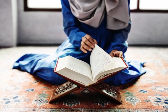 A Muslim woman in a blue dress and hijab reads a book on a patterned rug. The woman engages deeply with the book, showcasing a serene atmosphere of reading and reflection.