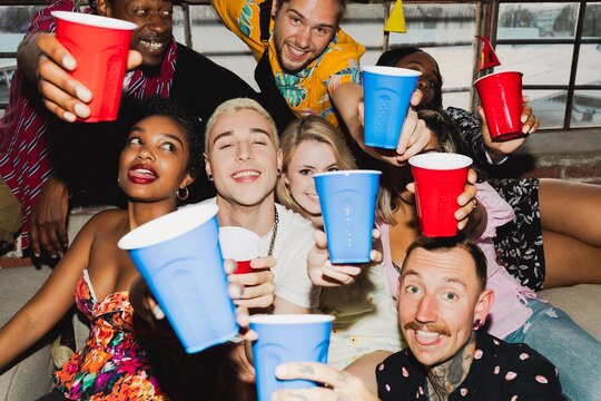 Group of diverse friends celebrating with red and blue cups, smiling and laughing. Mixed ethnicities, men and women, enjoying a fun party atmosphere. Diverse friends drinking at college dorm party.