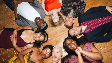 Diverse group of friends lying on wooden floor, smiling and laughing. Mixed ethnicities, men and women, enjoying a joyful moment together, surrounded by confetti. Diverse friends at festive home party