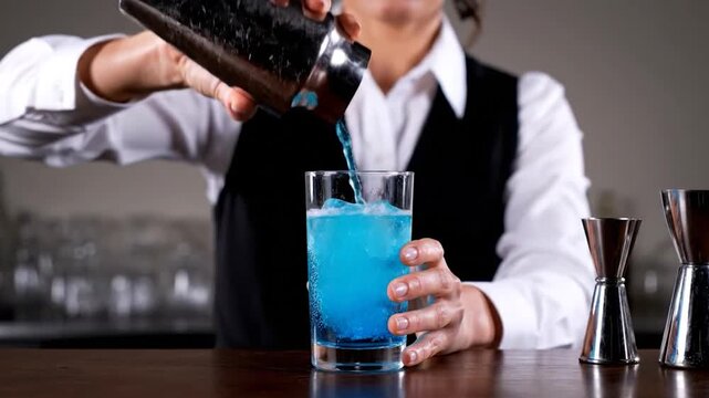 A bartender pours blue liquid from a shaker into a glass of ice, with bar tools nearby