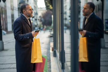 Mature man browsing shop window holding smartphone and shopping bags