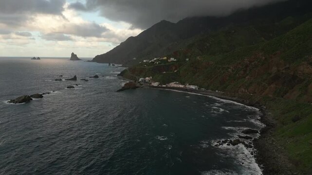 Coastal village nestled against verdant, mist-shrouded mountains. Rocky shores meet the dark, choppy sea under a dramatic sky.