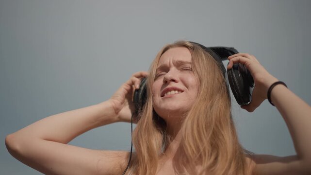 Caucasian woman with headset enjoying music, outdoor street setting swaying to beat while adjusting headset and smiling into sunlit sky, casual sleeveless top and beanie, long blonde hair moving