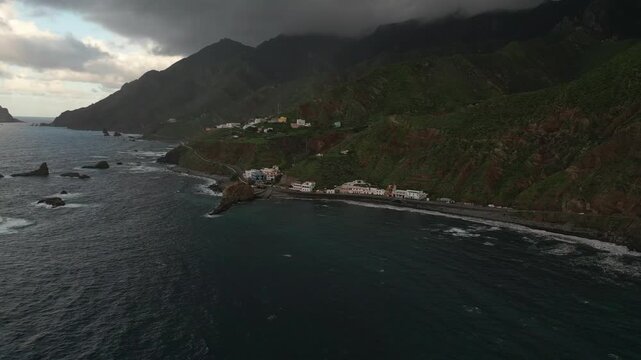 A coastal village nestles at the base of verdant, rugged mountains, facing a choppy, dark sea under a dramatic, cloudy sky.