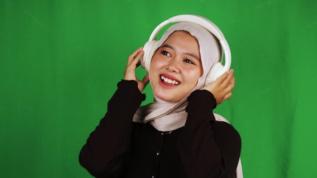 Excited Muslim woman in beige hijab wearing white headphones pointing up with idea gesture against green screen. Enthusiastic discovery moment perfect for music, podcast, inspiration, aha moment conte