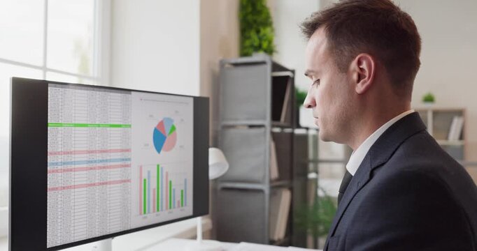 Accountant analyst auditor businessman at computer. Focused man in office reviews spreadsheet data on a monitor with charts and bars for finance report and audit. Audit finance concept in view.