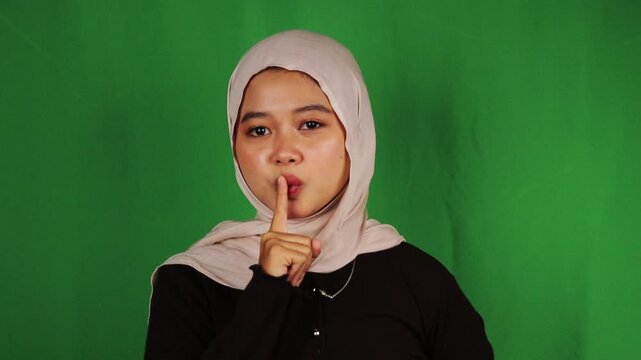 Muslim woman in beige hijab making shush gesture with finger on lips against green screen. Quiet signal perfect for silence, secret keeping, confidential, mystery, discretion, and library content.
