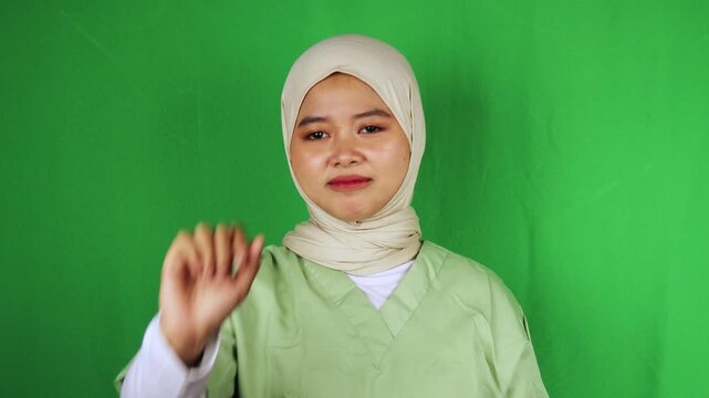 Muslim nurse in white hijab and green scrubs making stop hand gesture with awkward smile against green screen. Perfect for medical boundaries, patient care, healthcare communication, stop gesture, saf