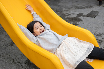 Excited Asian girl sliding quickly down a bright yellow playground slide with her arms raised, shouting with joy and adrenaline © Kwangmoozaa