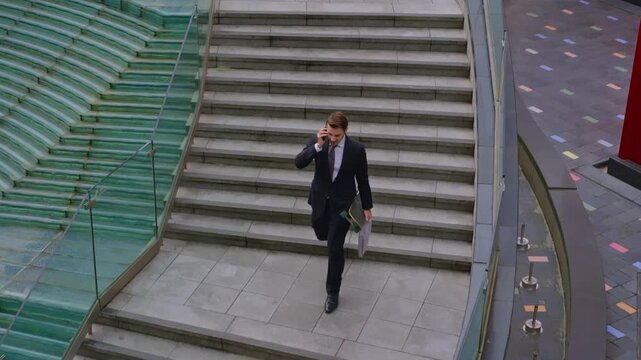 High angle tracking shot of young businessman in suit talking on smartphone while rushing down stairs carrying skateboard and laptop bag in modern urban environment
