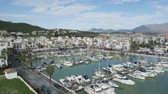 Drone footage of Puerto de la Duquesa in Manilva, showing boats, waterfront apartments, hills, and the Mediterranean coastline.