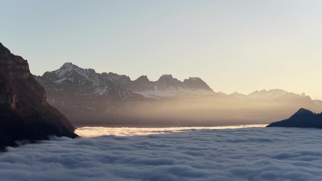 Aerial of the Swiss Alps. Sunbeams piercing through a thick sea of fog in the Walensee valley, illuminating the jagged peaks of Churfirsten in a warm, cinematic winter glow.