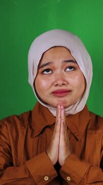 Grateful Muslim woman in white hijab and brown shirt with praying hands looking up joyfully against green screen. Thankful prayer perfect for gratitude, blessing, faith, worship, spiritual, religious 