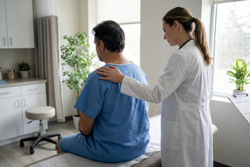 Fototapeta premium A female doctor places a reassuring hand on the back of a middle-aged male patient wearing a blue gown sitting on an exam table in a bright medical clinic room.