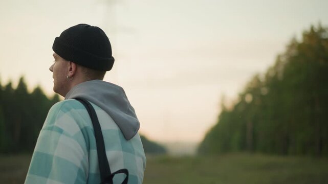 wide shot caucasian backpacker gazing sunset horizon, pensive young man in beanie and plaid jacket with backpack straps, open meadow and pine line, soft twilight glow, contemplative mood of discovery