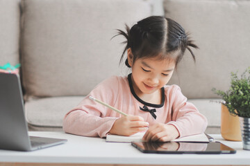 Naklejka premium A cheerful Asian mother helps her young daughter do homework and color a book at home, while the father works on a laptop in the background. A cozy, bright, and modern family lifestyle.