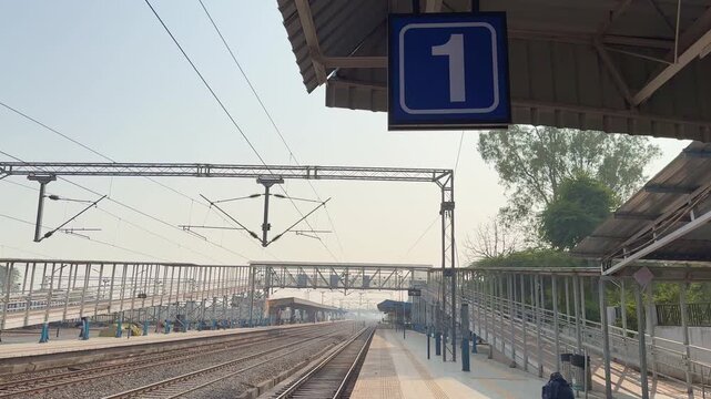 Tilt up shot of platform number one sign with railway tracks, overhead electric lines and foot overbridge visible under clear sky at an Indian railway station