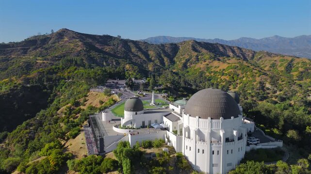 Aerial view of Griffith Observatory surrounded by green hills and mountains above Los Angeles California, iconic landmark overlooking the city skyline under clear blue sky
