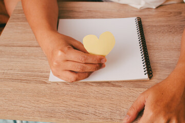 Close up of hand holding a yellow paper heart over wooden table, concept of kindness, romantic message, and self-love support.