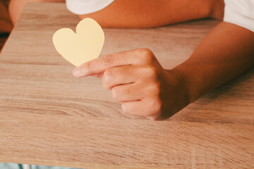 Close up of hand holding a yellow paper heart over wooden table, concept of kindness, romantic message, and self-love support.