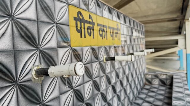 Drinking water taps mounted on patterned tiled wall at railway platform with Hindi sign, clean public water facility for passengers inside covered station area daylight