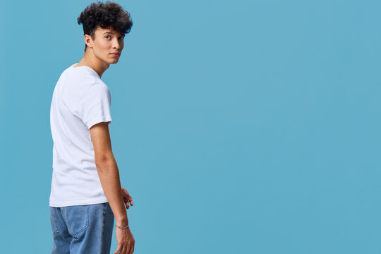 young man in casual white t-shirt and blue jeans with curly hair looking back against plain blue background, modern style, isolated, neutral expression, studio portrait
