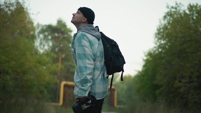 white male scout walking under pipeline, backpack and camera gear on shoulder, scouting shoot location where industrial pipe meets green foliage, overgrown trail, cinematic framing and contemplative
