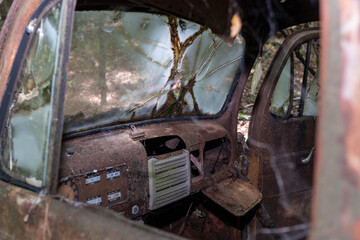 Fototapeta premium Rusted, abandoned truck interior exposed to the elements, featuring a shattered windshield, corroded dashboard, and overgrown surroundings. Evokes decay, history, and the passage of time.
