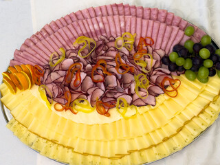 Cold cuts and cheese platter arranged on a serving tray with sliced ham, smoked meat, cheese, grapes and vegetables. Catering food presentation suitable for buffet, celebration or event.