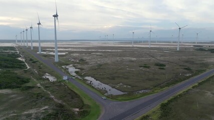 Aerial drone pull back rising reveal view of road intersection with car driving near wind farm turbines over wetlands infrastructure in Tramandai Brazil. 4K 60fps 10-bit D-Log video.