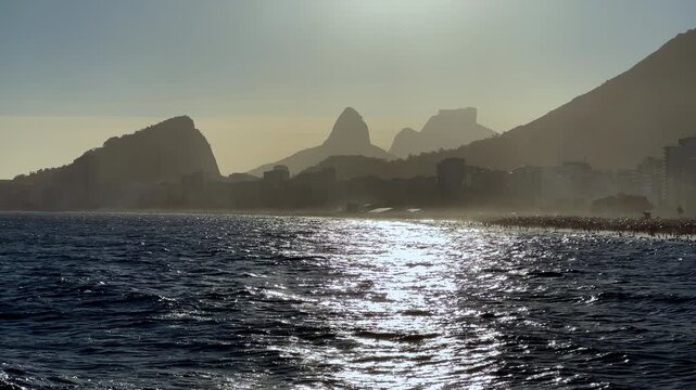 Cinematic view of the sunset reflection on the sea in Rio de Janeiro. Features the golden light shimmering on the waves with the silhouettes of Sugarloaf Mountain and the Copacabana skyline in the bac