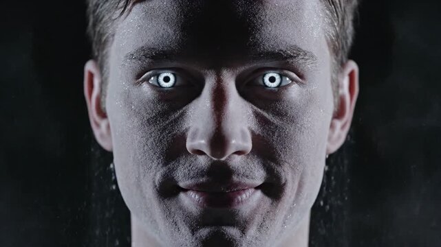Closeup of a mans face covered in white powder or mist with eyes closed against a dark background