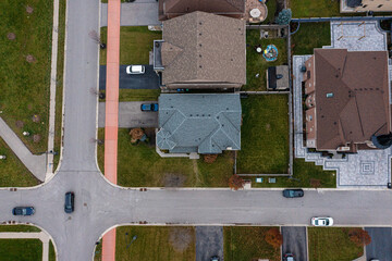 Overhead drone view showing a planned suburban neighborhood with houses, green lawns, asphalt roads, sidewalks, and cars, depicting modern residential living and urban development