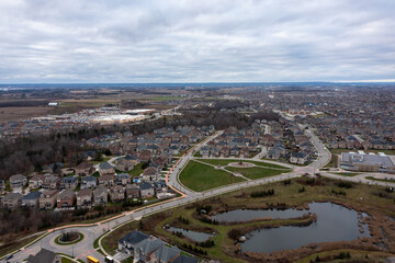 Fototapeta premium Aerial view showing suburban residential development with many houses. Streets. A roundabout. And retention ponds contrasting against open agricultural land and a commercial plaza in brampton. Ontario