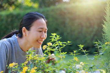 Close-up of a happy Asian woman smiling and enjoying purple flowers in her green garden. Beautiful female portrait in sunny backyard park. Concept of nature lovers, wellness, and outdoor lifestyle.