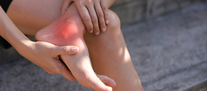 Close-up of woman hand holding painful injured ankle with red inflamed spot and bruising. Medical concept of joint pain, sprain, ligament injury, bone ache, or arthritis in outdoor background.
